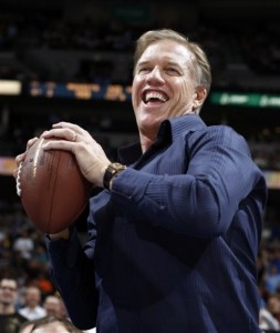 In this Feb. 9, 2010, file photo, retired Denver Broncos quarterback John Elway prepares to throw a football to Denver Nuggets mascot Rocky the Mountain Lion in the third quarter of the Nuggets' 127-91 victory over the Dallas Mavericks in an NBA basketball game in Denver. Elway, who retired in 1999 after winning his second straight Super Bowl in Denver, is planning to attend some of the Broncos' training camp practices this summer and has agreed to help market the team's game in London in September.  (AP Photo/David Zalubowski,File)